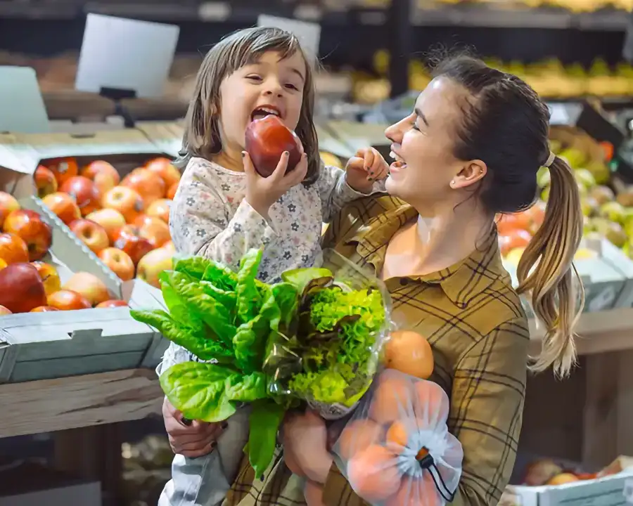 Smiling parent holding a child biting into a red apple in the produce aisle at Food Fantasies near Jacksonville, IL